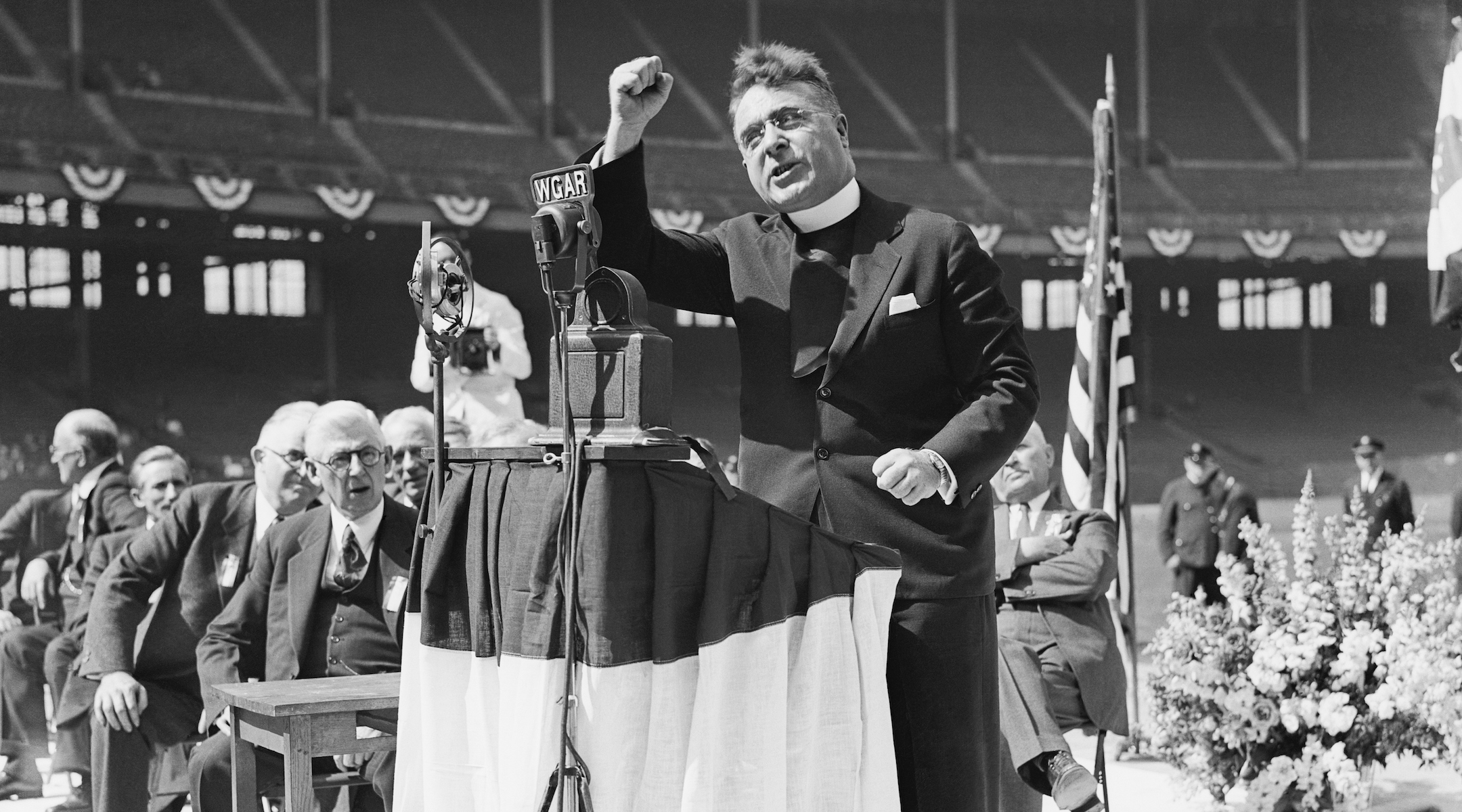 A black-and-white photo of a priest shaking his fist at a lectern at an outdoor rally.